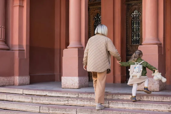 Back view of woman and child with teddy bear holding hands while walking on stairs near building — Stock Photo