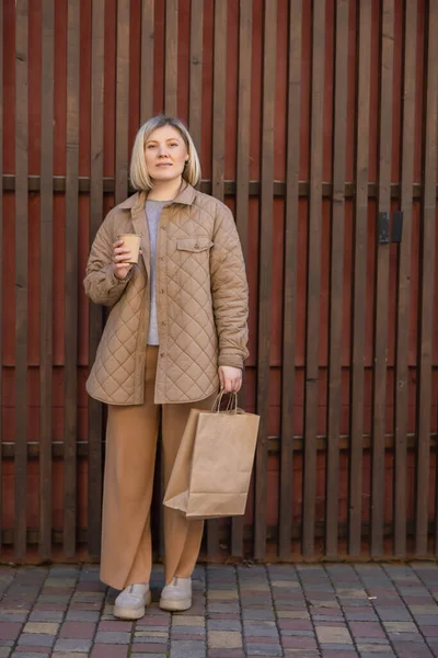 Full length view of trendy woman with coffee to go and shopping bag looking at camera near wooden fence — стоковое фото