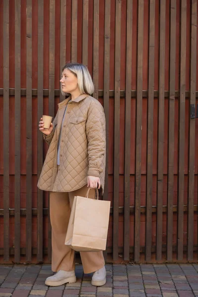 Full length view of stylish woman standing with paper cup and shopping bag near wooden fence — Foto stock