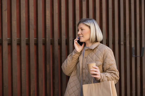 Mujer rubia sonriente con café para ir y bolsa de compras hablando en el teléfono inteligente cerca de valla - foto de stock