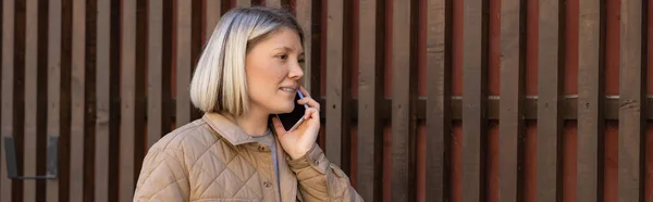 Smiling blonde woman talking on smartphone near wooden fence, banner — Photo de stock