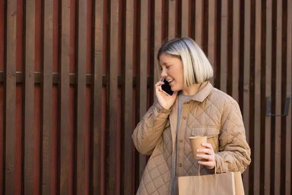 Happy woman in beige jacket talking on mobile phone while holding paper cup and shopping bag — Stockfoto