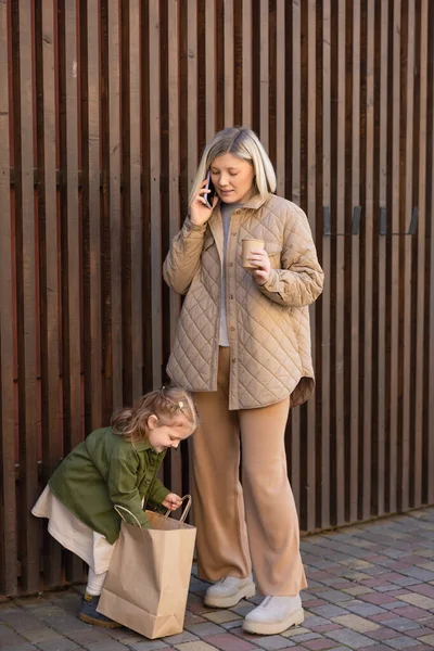 Mujer sonriente con bebida para llevar hablando en el teléfono celular cerca de la hija mirando en la bolsa de compras - foto de stock