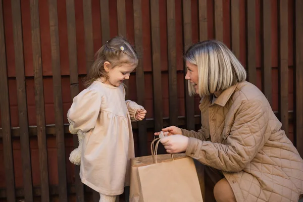 Happy woman showing shopping bags to little daughter near wooden fence — Fotografia de Stock