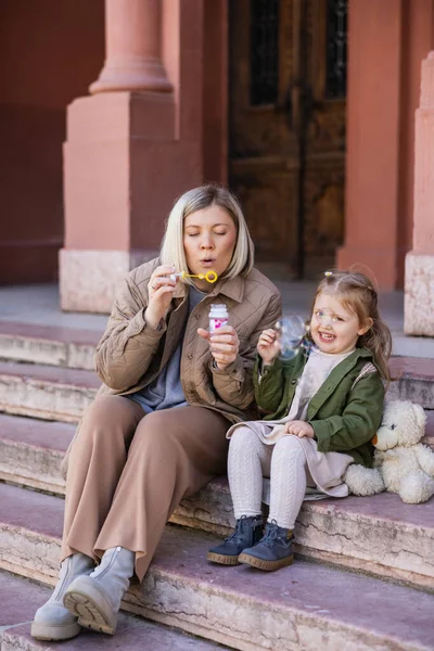 Woman blowing soap bubbles near happy daughter on stairs outdoors — Stock Photo