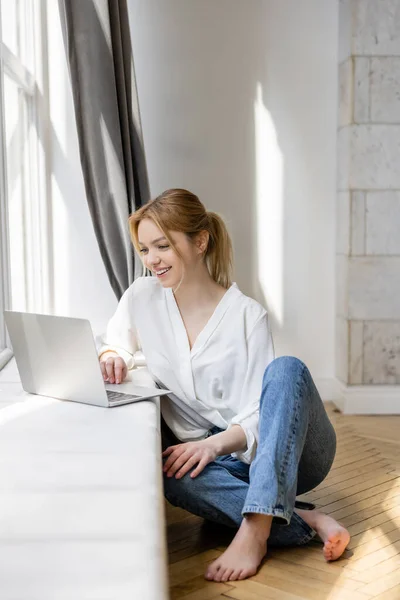 Freelancer sorridente usando laptop no peitoril da janela em casa — Fotografia de Stock