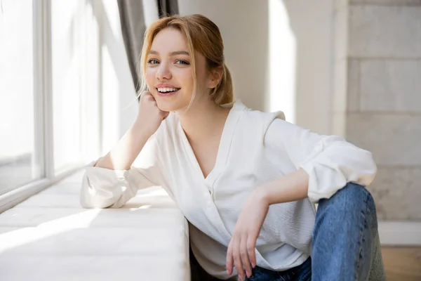 Portrait of smiling blonde woman looking at camera near windowsill at home — Stock Photo