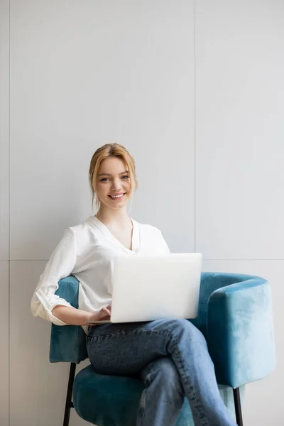 Positive freelancer holding laptop and looking at camera while sitting on armchair at home — Stock Photo