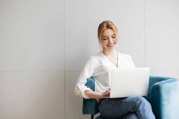 Positive blonde Frau benutzt Laptop, während sie zu Hause auf einem Sessel sitzt — Stockfoto