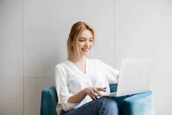 Glückliche junge Freiberuflerin in Bluse mit Laptop auf Sessel zu Hause — Stockfoto