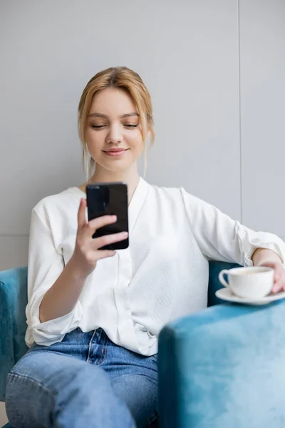 Positive blonde Frau benutzt verschwommenes Smartphone und hält Tasse auf Sessel zu Hause — Stockfoto