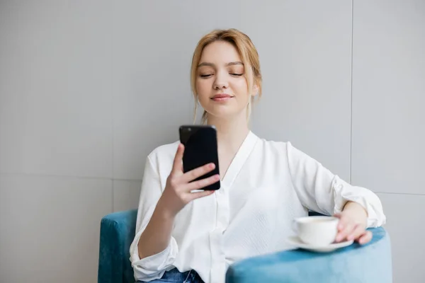 Hübsche blonde Frau benutzt Smartphone in der Nähe verschwommener Kaffeetasse auf Sessel — Stockfoto