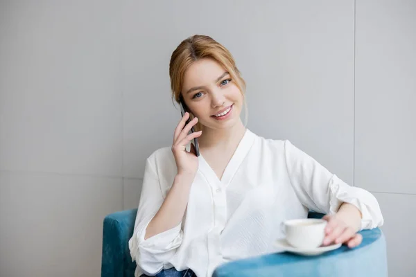 Glückliche junge Frau spricht auf Smartphone und hält verschwommene Tasse auf Sessel — Stockfoto