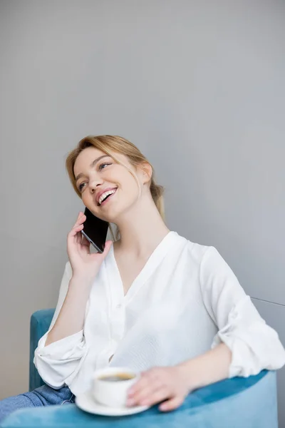 Mujer joven positiva hablando en el teléfono celular cerca de taza borrosa de café en el sillón - foto de stock
