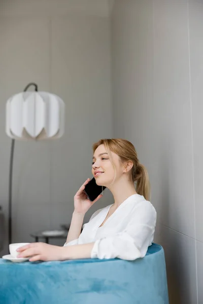 Mujer sonriente en blusa hablando en teléfono inteligente y sosteniendo la taza en el sillón - foto de stock