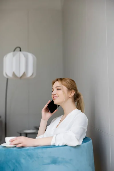 Femme souriante en chemisier parlant sur smartphone et tenant la tasse sur fauteuil — Photo de stock