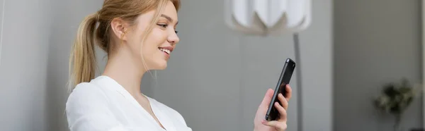 Jeune femme positive en chemisier utilisant un téléphone portable à la maison, bannière — Photo de stock