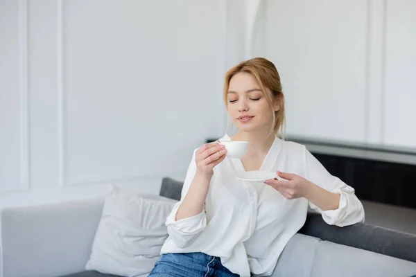 Pretty blonde woman holding cup and saucer on couch at home — Stock Photo