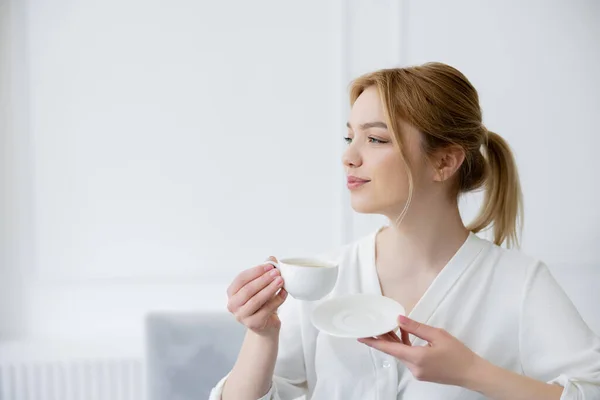Jolie jeune femme tenant tasse de café et soucoupe à la maison — Photo de stock