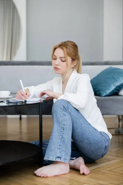 Barefoot woman writing on notebook near coffee on table at home — Stock Photo