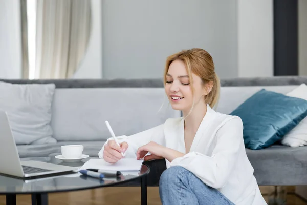 Smiling freelancer writing on notebook near laptop and coffee at home — Stock Photo