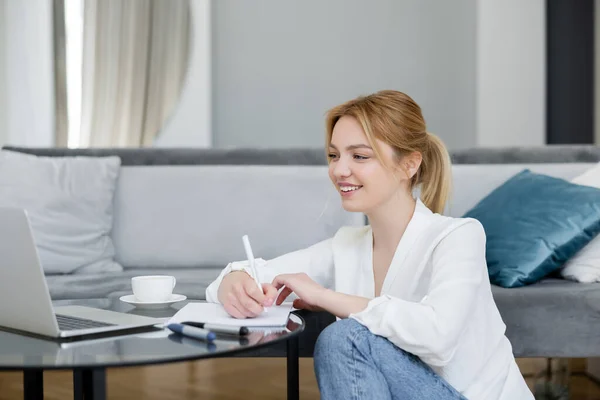 Positive young freelancer writing on notebook near laptop on coffee table at home — Stock Photo