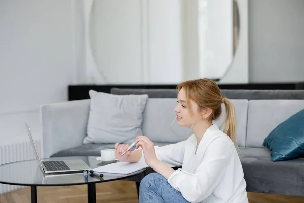 Junge Freiberuflerin schaut zu Hause auf Laptop und Notizbuch — Stockfoto