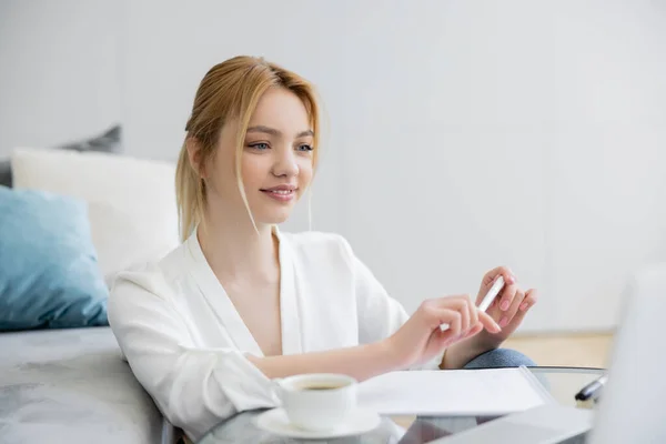 Hübsche Freiberuflerin schaut zu Hause auf verschwommenen Laptop neben Tasse und Notizbuch — Stockfoto