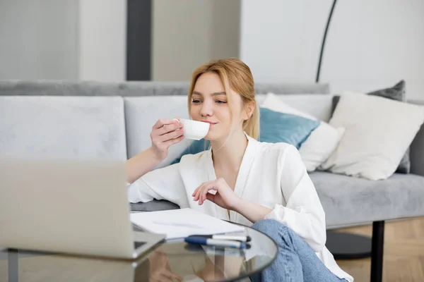 Freelancer en blusa sosteniendo taza cerca de laptop y notebook en sala de estar - foto de stock