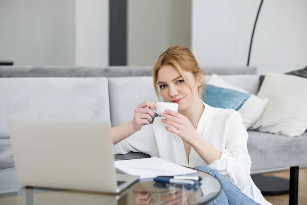 Blonde Freiberuflerin hält Tasse neben verschwommenem Laptop und Notizbuch auf Wohnzimmertisch — Stockfoto
