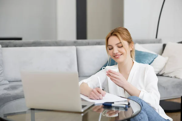 Cheerful freelancer looking at coffee cup while writing on notebook near laptop in living room — Stock Photo