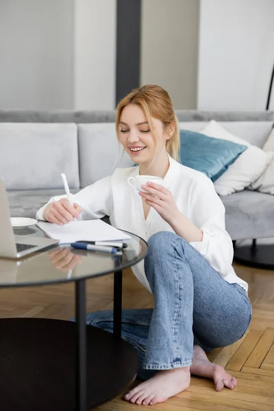 Pretty blonde freelancer holding cup and writing on notebook near laptop on coffee table in living room — Stock Photo