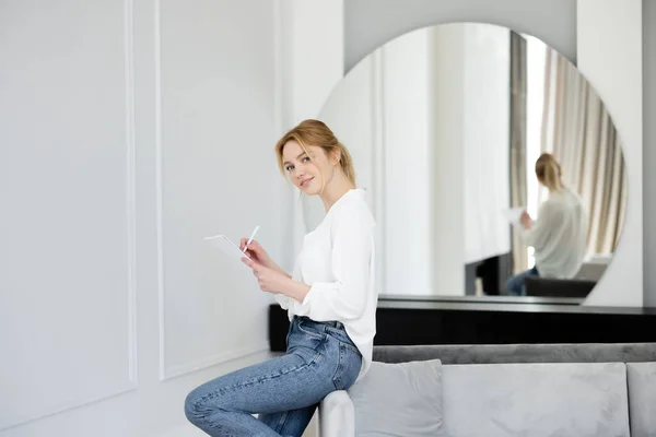 Hübsche junge Frau schreibt auf Notizbuch und blickt im Wohnzimmer in die Kamera — Stockfoto