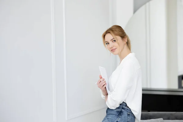 Glückliche junge Frau hält Notizbuch in der Hand und blickt im Wohnzimmer in die Kamera — Stockfoto