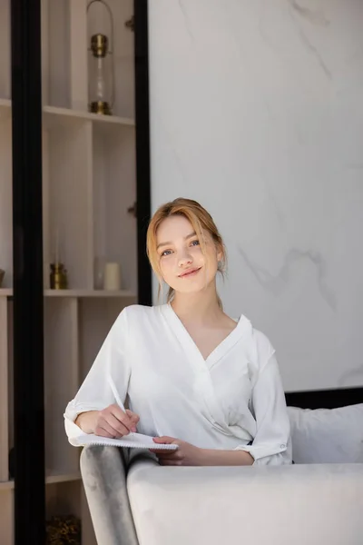 Hübsche Frau in Bluse schreibt auf Notizbuch auf Couch zu Hause — Stockfoto