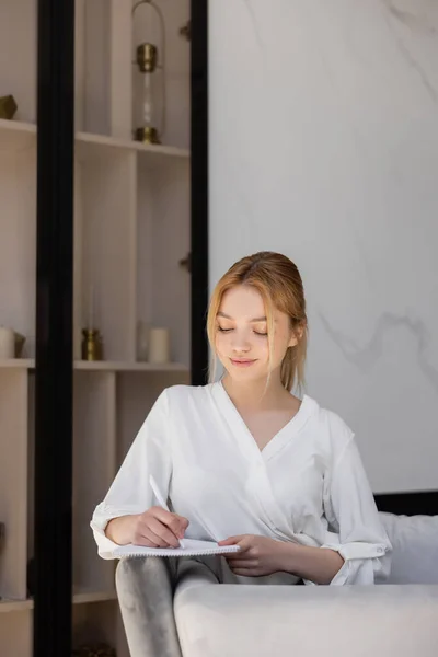 Smiling blonde woman writing on notebook on couch at home — Stock Photo