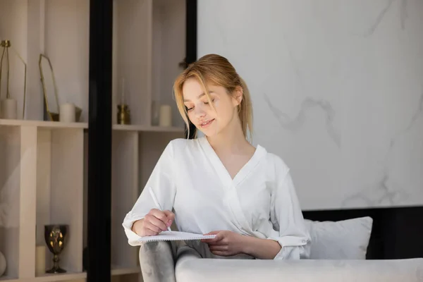 Junge Frau in Bluse schreibt auf Notizbuch auf Couch im Wohnzimmer — Stockfoto