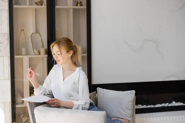 Junge Frau in weißer Bluse hält Notizbuch und Stift zu Hause auf Sessel — Stockfoto