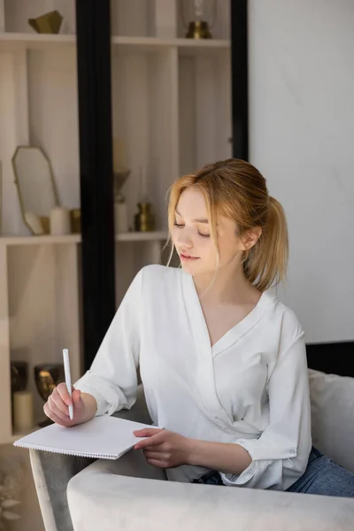 Hübsche junge Frau schreibt auf Notizbuch, während sie zu Hause im Sessel sitzt — Stockfoto