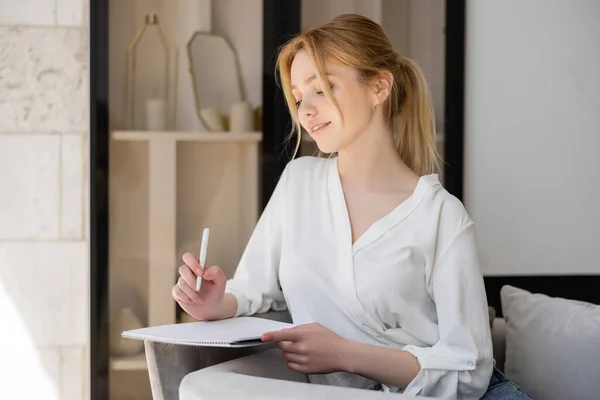 Mulher loira de blusa segurando caneta e caderno em casa — Fotografia de Stock