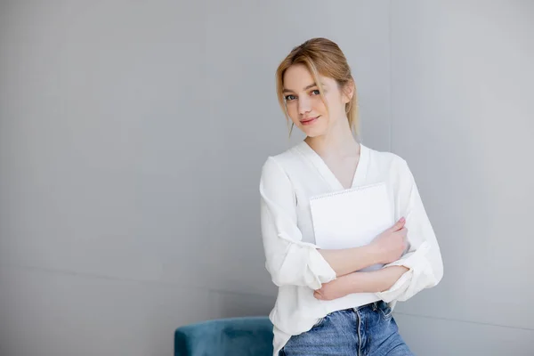 Blonde woman in white blouse looking at camera and holding notebook near armchair at home — Stock Photo