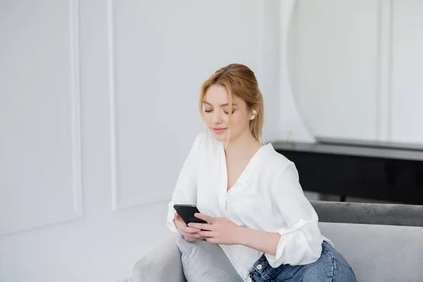 Young woman using smartphone and earphone on couch at home — Stock Photo