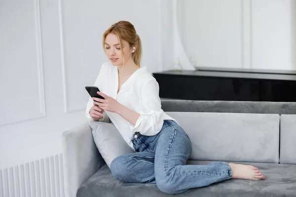 Barefoot woman in earphone using mobile phone on couch at home — Stock Photo