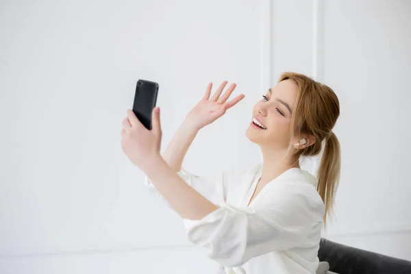 Positive blonde woman in earphone having video call on cellphone at home — Stock Photo