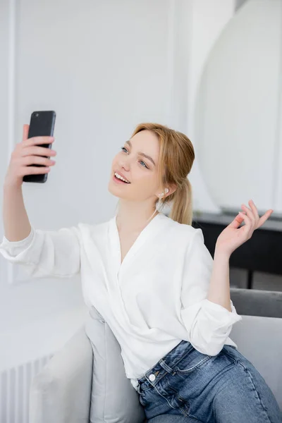 Mujer joven positiva en blusa y auricular que tiene videollamada en teléfono inteligente en la sala de estar - foto de stock