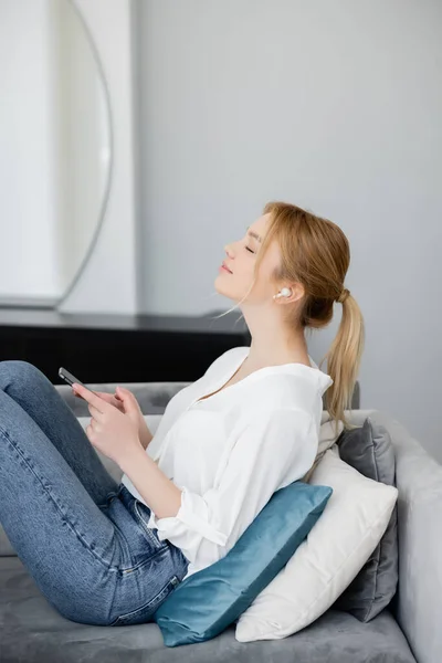 Vista lateral da jovem mulher ouvindo música no fone de ouvido e segurando smartphone no sofá — Fotografia de Stock