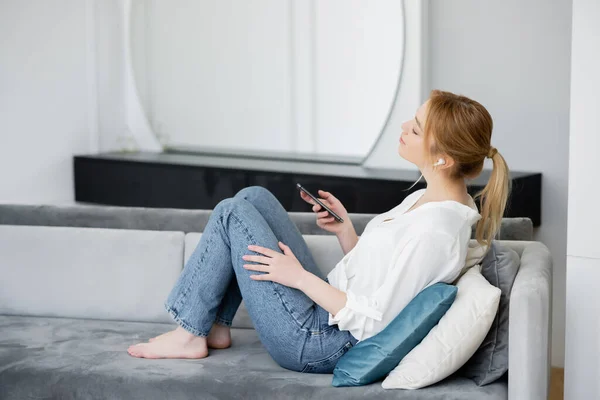 Vista lateral de una mujer joven en auricular sosteniendo el teléfono celular en el sofá en casa - foto de stock