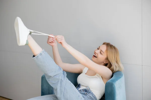 Mujer joven positiva sosteniendo el cordón de zapatilla de deporte mientras está sentado en el sillón - foto de stock