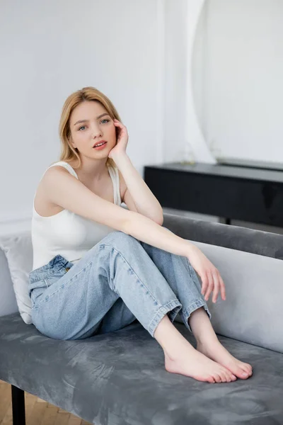 Barfüßige Frau in Jeans blickt in die Kamera, während sie zu Hause auf der Couch sitzt — Stockfoto