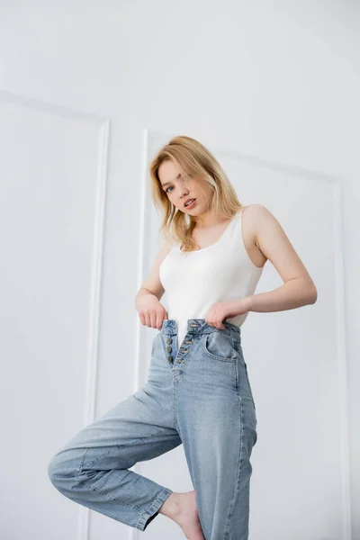 Mujer joven en body tocando jeans y mirando a la cámara en la sala de estar - foto de stock